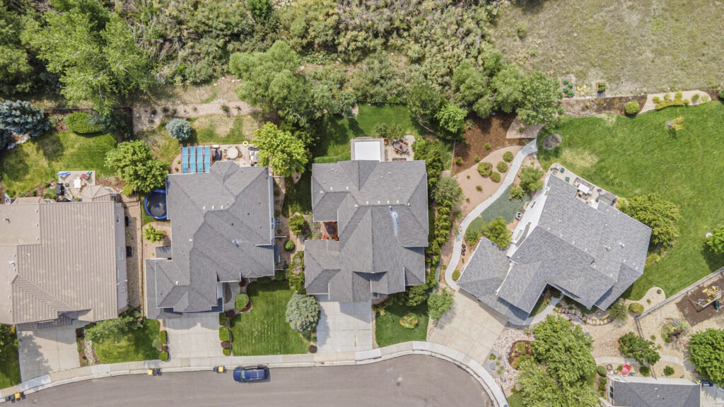 Bird’s-eye aerial drone view of a modern luxury two-story home featuring a three-car garage, professional landscaping, and lush vegetation behind the home for added privacy.