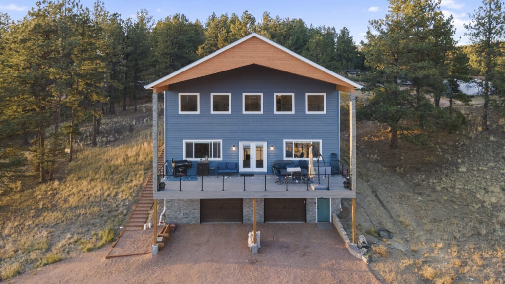 Aerial drone photo of the exterior of a newly built two-story mountain home in Colorado featuring a large deck, two-car garage, and abundant windows.