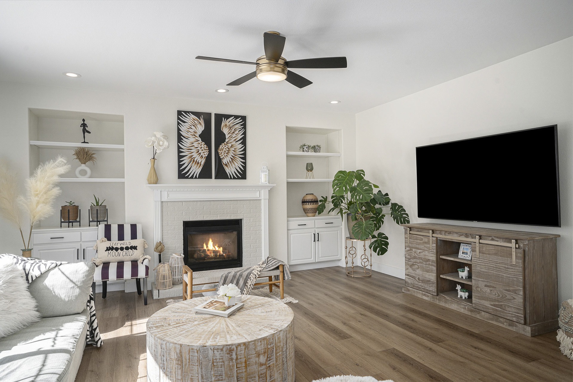 Bright living room with natural wood floors, a ceiling fan, large windows for natural light, and a fireplace.